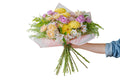 A hand holding a vibrant floral bouquet with yellow chrysanthemums, pink roses, and white alstroemeria wrapped in light pink tissue paper, set against a solid light gray background.